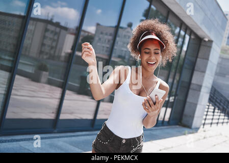 Junge Frau mit Tennis Visor und earphones Free Style auf der Straße zu Fuß in der Nähe der Fenster holding Smartphone hören Lieblingslied singen smil Stockfoto