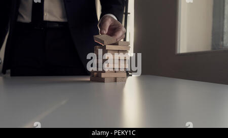 Geschäftsmann in einem schattigen Büro platzieren Holzklotz auf Turm mit einem natürlichem Sonnenlicht von hinten. Stockfoto