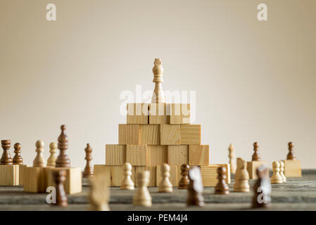 Weiße König auf hölzernen Bausteinen mit anderen organisiert verschiedene Schachfiguren unter der Pyramide. Stockfoto