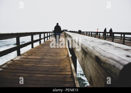 Steg Bridge, verschwommener Hintergrund von Menschen, die an einem bewölkten Tag mit Sturm auf dem Holzsteg in Swakopmund an der Küste Westnamibias Afrikas spazieren gehen. Stockfoto