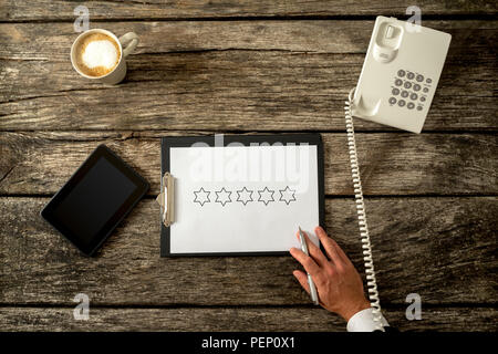 Unternehmer im Gespräch über ein Telefon mit fünf Sterne für Qualität sichtbar auf einem Notizblock auf einem Klemmbrett auf einem rustikalen Holzmöbeln, Schreibtisch oder Tisch mit einer Tablette alongsid Stockfoto