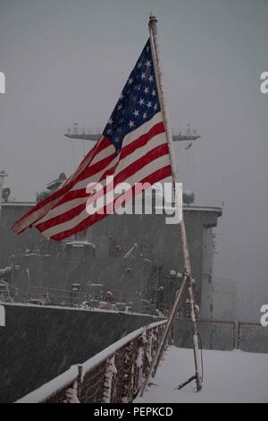 Am 24. Januar 2016, an Bord der USS Ashland (LSD 48) in Sasebo, Japan, flattert die Fähnrich auf dem schneebedeckten Flugdeck, bevor die Abendfarben erscheinen. Das amphibische Dock-Landungsschiff, Teil der Bonhomme Richard Amphibious Ready Group, ist dabei, die geplante Continuous Maintenance Availability (CMAV) abzuschließen. Die Seeleute sind aufmerksam, während die Zeremonie den Respekt für das nationale Fähnrich und die Einsatzbereitschaft bei routinemäßigen Instandhaltungs- und Ausbildungseinsätzen kennzeichnet. Stockfoto