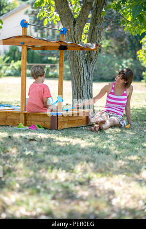 Junge Mutter spielt mit ihren Toddler Sohn saß in einer Sandbox unter einem Baum an einem sonnigen Sommertag. Stockfoto