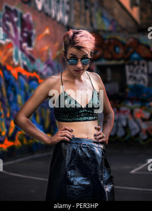 Street Punk oder Hippie Mädchen mit rosa gefärbte Haare. Frau mit Piercing in der Nase, Ohren Tunnel und ungewöhnliche Frisur am Basketballplatz mit Graffiti pai Stockfoto