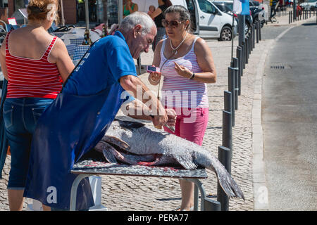 Setubal, Portugal - August 8, 2018: Mann, der die Waage der ein grosser Fisch in der Straße entfernt vor einem Restaurant an einem Sommertag unter den Augen von Stockfoto