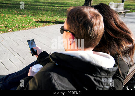 Paar sitzen auf einer Bank im Stadtpark, ein junger Mann hält einen Kaffee trinken gehen und zeigt etwas zu dem Mädchen auf Smartphone und genießen die Sonne. Stockfoto