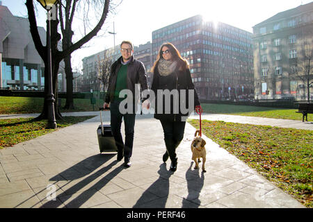 Paar mit Koffer und Hund Fuß in den Park am frühen Morgen. Glückliches junges Paar zu Fuß durch den Park mit niedlichen Hund und genießen. Stockfoto