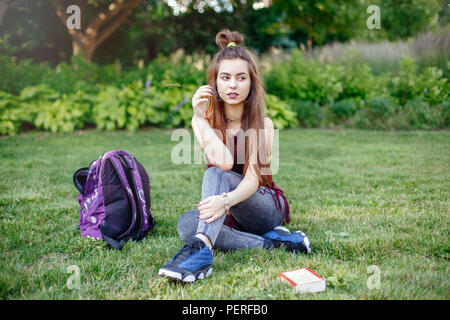 Portrait von schöne nachdenkliche weiße Kaukasier junge Frau Schüler weiblich, sitzend auf Gras im Park außerhalb mit Rucksack und Buch. Hobby Bildung ba Stockfoto