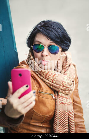 Portrait von mittleren Alters kaukasischen brünette Frau mit kurzen Bob Haar in Sonnenbrille trägt Hellbraun leder jacke und Schal außerhalb machen selfie Stockfoto