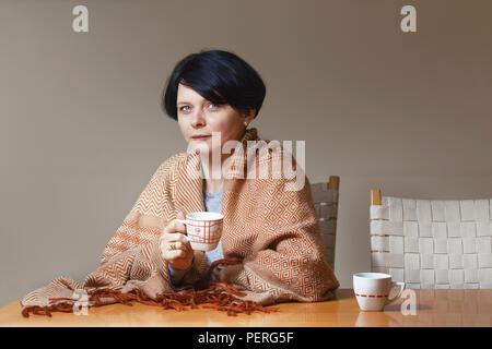 Porträt der schönen kaukasischen Brünette Mitte Alter Frau mit Decke am Tisch trinken Tee Kaffee Tasse in den Händen halten, sitzen direkt abgedeckt Stockfoto