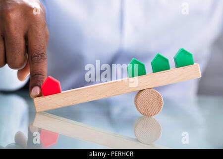Menschliche Hand Balancing Rote und Grüne Miniatur Haus auf Holz- Wippe über die reflektierende Schreibtisch Stockfoto