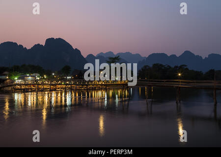 Silhouette einer karst Kalkstein Berge, Brücke und wenige Leute an einem beleuchteten Waterfront Restaurant am Nam Song Fluss in Vang Vieng, Laos, bei Sonnenuntergang. Stockfoto