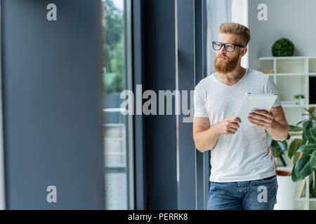 Arbeiten an neuen Tablet. Low Angle View der selbstbewussten jungen Mann bei der Arbeit mit digitalen Tablet beim Stehen vor dem großen Fenster im Büro oder Cafe. Stockfoto