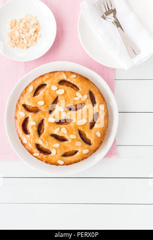 Hausgemachte Victoria Pflaumenkuchen auf einem weißen Kuchen Platte, eine kleine Platte mit gerösteten Mandeln, Platten und Kuchen Gabeln auf weißem Holz Planken. Stockfoto