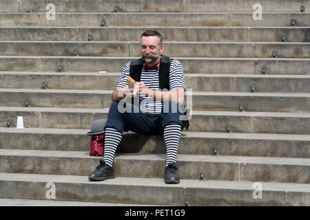 Freudiger Mann mittleren Alters in gestreifter Clown Kostüm sitzt auf der Treppe der Vorbereitung einer sandvich zu essen Stockfoto