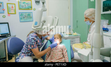 Kind mit einer Mutter ein Empfang der Zahnarzt. Das Mädchen liegt in der Stuhl, hinter ihrer Mutter. Der Arzt arbeitet mit einem Assistenten. Verfahren für das Bohren eines Zahnes. Die Einsetzung eines nichtständigen Dichtung. Stockfoto