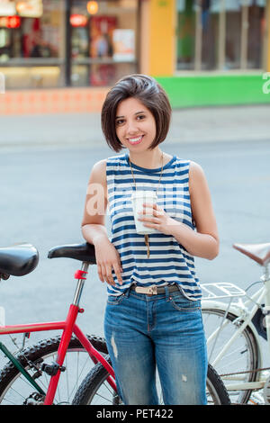 Porträt der Schönen lächelnden Jungen latin kolumbianische Mädchen Frau mit kurzen Haaren Bob in Blau zerrissene Jeans gestreiftes T-Shirt, Tasse Kaffee schiefen o Stockfoto