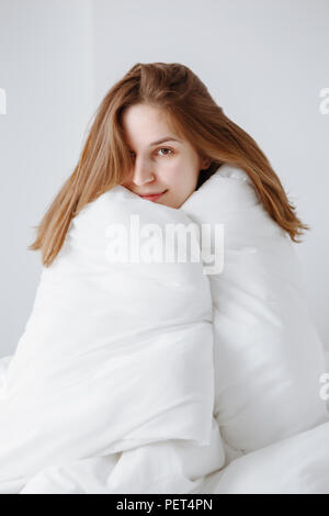 Portrait von lächelnden schönen jungen kaukasischen Mädchen Frau mit langen roten Haaren im Bett sitzend mit Decke gewickelt auf weißem Hintergrund, suchen Stockfoto