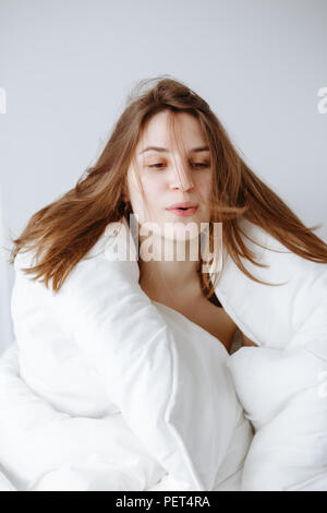 Portrait von schönen jungen kaukasischen Mädchen Frau im Bett sitzend mit Decke gewickelt auf weißem Hintergrund, ihr Haar weht am frühen Morgen, l Stockfoto