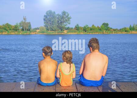 Kinder sitzen auf Pier. Drei Kinder verschiedenen Alters - jugendlicher Junge, elementare alter Junge und Mädchen sitzen auf einem hölzernen Pier. Sommer und Kindheit Konzept. Kinder auf der Bank am See. Geschwister sitzen auf hölzernen Pier Stockfoto