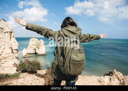 Ansicht von der Rückseite. Eine junge Frau tourist genießt die schöne Aussicht auf den Atlantischen Ozean und die Landschaft an der Küste in Portugal und hebt die Arme nach oben zeigen, wie froh sie ist. Stockfoto