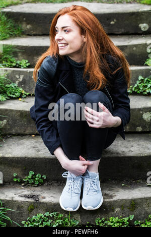 Portrait von lächelnden jungen Frau mit langen roten Haaren, sitzen auf Schritte im Freien. Stockfoto