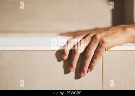 Nahaufnahme der Hand eines jungen kaukasischen Mann auf der Seite der weißen Wanne, während es in der Badewanne, in einem beigefarbenen Badezimmer Stockfoto