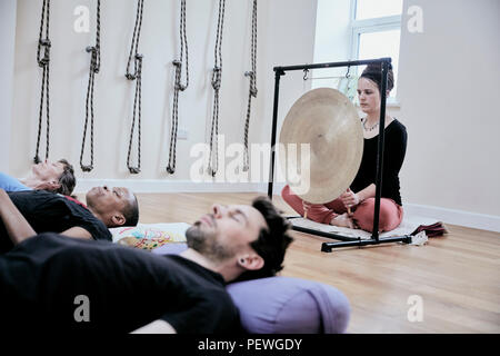 Frau mit einem Gong während einer klangtherapie Sitzung und drei Menschen auf dem Rücken liegend entspannen. Stockfoto