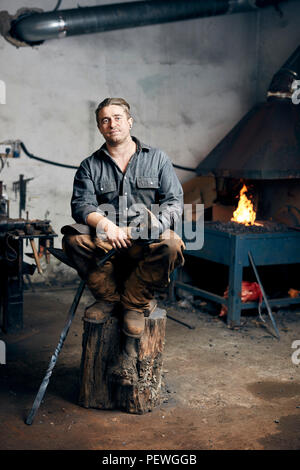 Porträt eines handwerklichen Schmied in seiner Werkstatt, sitzen auf einem Holzklotz Holding eine lange Hammer. Stockfoto