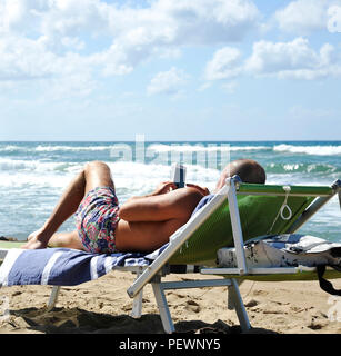 Mann am Strand liegend auf der Sonnenbank auf das Smartphone. Im Hintergrund zwei Männer das Meer betrachten. Es ist Kontemplation und contemplatio Stockfoto