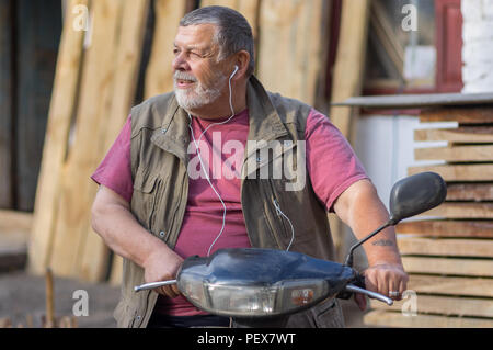 Schöne Outdoor Portrait von bärtigen älteren Mann sitzt auf einem Roller und Musik hören Stockfoto