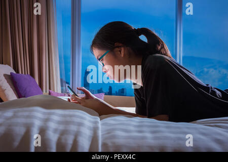 Asiatische jugendlicher Mädchen mit Ihrem Smart Phone auf soziale Medien allein in ihrem Schlafzimmer mit einem großen Fenster mit Blick auf die Stadt am späten Abend anschließen, gut für Modus Stockfoto