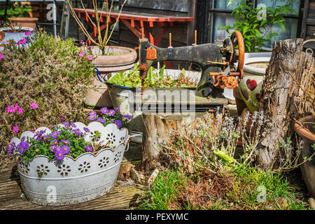 Eine alte Nähmaschine der Marke Phoenix steht auf einem Baumstamm. Es gibt auch viele bunte Blumen. Stockfoto