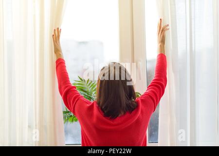 Frau schaut aus dem Fenster, und öffnet die Gardinen. Ansicht von der Rückseite Stockfoto