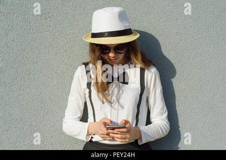 Outdoor Portrait von Jugendmädchen in Hut, Mädchen verwendet, Smartphone, liest, schreibt Nachricht, grau texturierte Wand Hintergrund Stockfoto