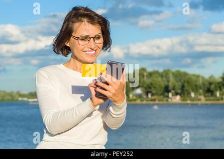 Nach Frau sieht Visitenkarte und macht einen Anruf. City Park Hintergrund, Fluss, Himmel mit Wolken. Stockfoto