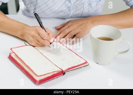Die Hand einer Frau mit einem Stift schreibt in ihrem Tagebuch. Weiter auf dem Tisch ist eine Tasse Kaffee. Stockfoto