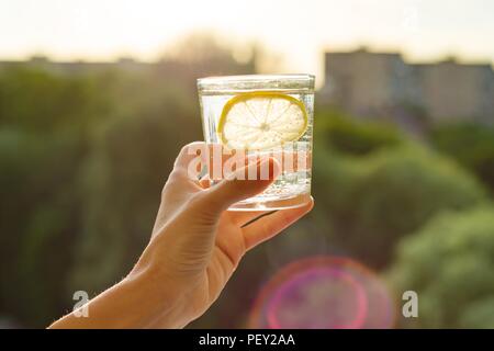 Glas klar, Mineralwasser mit Zitrone in der Hand. Hintergrund Himmel, Silhouette der Stadt, Sonnenuntergang. Stockfoto
