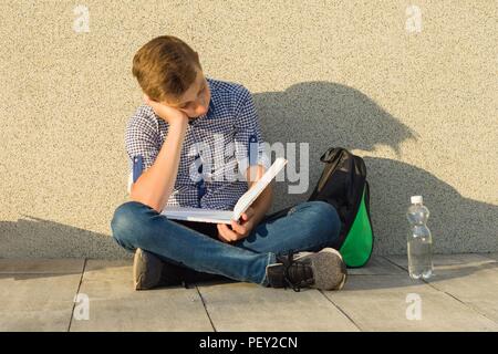 Jugendlicher Schüler liest Lehrbuch, sitzt in der Nähe der graue Wand der Schule Stockfoto