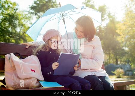 Mutter und Tochter gemeinsam auf einer Bank im Stadtpark ruht, das Mädchen in der Brille liest, zu ihrer Mutter eine Schule Notebook, und spricht über die Schule l Stockfoto