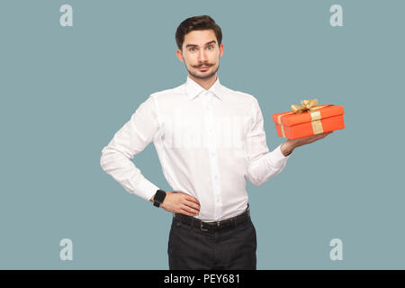 Happy bärtiger Mann im weißen Hemd stehend mit der Hand an der Hüfte halten roten Geschenkbox, an Kamera mit zufriedenen Gesicht und lächelt. Innen-, Studio s Stockfoto