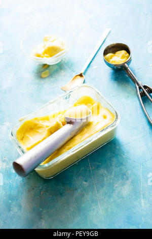 Mango Eis auf einem Glasbehälter mit Icecream Utensilien, auf einem blauen Brett, frontale Ansicht mit natürlichen Licht zurück Stockfoto