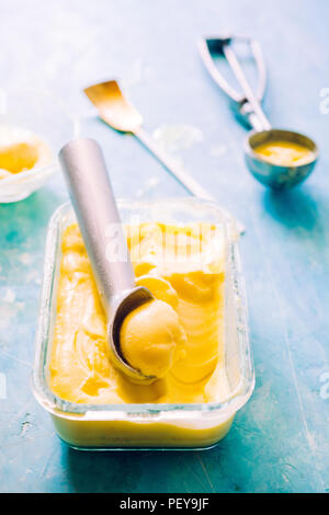 Mango Eis auf einem Glasbehälter mit Icecream Utensilien, auf einem blauen Brett, frontale Ansicht mit natürlichen Licht zurück Stockfoto