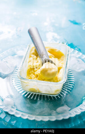 Mango Eis auf einem Glasbehälter mit Icecream Utensilien, auf einem blauen Brett, frontale Ansicht mit natürlichen Licht zurück Stockfoto