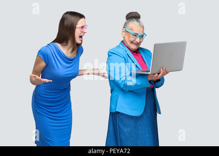 Unglückliche junge Frau im blauen Kleid schrie gealterte Frau Arbeiten am Laptop. Beziehungen oder Beziehung, die auf der Familie zwischen Enkelin und Oma. Stockfoto