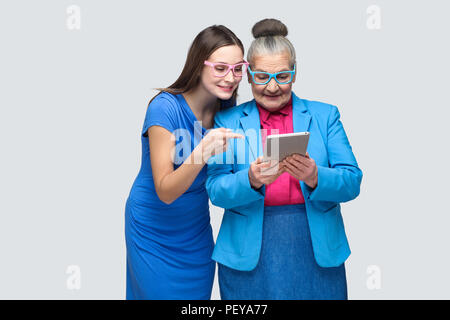 Junge Tochter in blauem Kleid zeigen, zeigen mit dem Finger auf Tablet Display in die Hände der gealterte Frau. Familie Beziehungen zwischen Enkelin und Oma Stockfoto