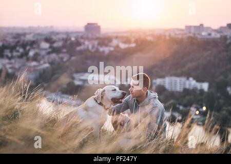 Morgen Spaziergang mit Hund. Junger Mann und seinem Labrador Retriever auf der Wiese gegen Stadt bei Sonnenaufgang. Stockfoto