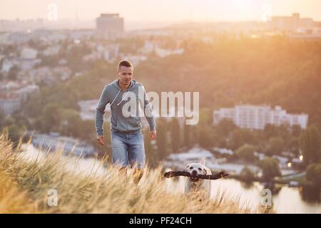 Morgen Spaziergang mit Hund. Junger Mann und seinem Labrador Retriever auf der Wiese gegen Stadt bei Sonnenaufgang. Stockfoto