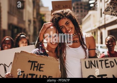 Porträt von zwei glückliche junge Frau protestieren mit Plakaten im Außenbereich mit einer Gruppe von Menschen im Hintergrund. Stockfoto