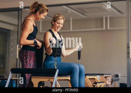 Lächelnde Frau an der Turnhalle tun Pilates Training mit Ihrem Trainer. Frau ziehen Strech Bänder mit beiden Händen sitzen auf Pilates Training Maschine. Stockfoto